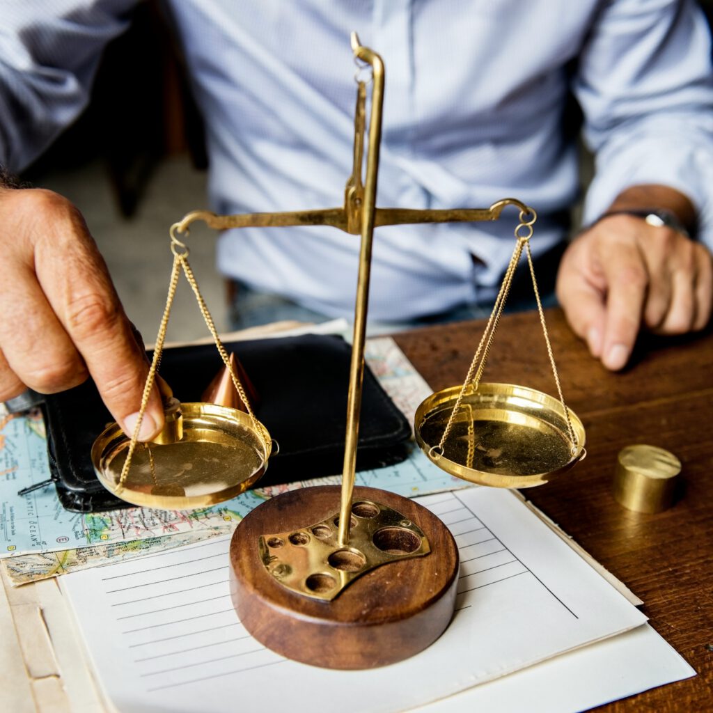 Person justiert eine goldene Waage auf einem Schreibtisch mit Dokumenten, Landkarte und Schreibutensilien – Symbol für Gerechtigkeit und Ausgewogenheit