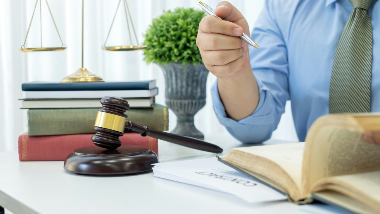 Ein Anwalt oder Richter sitzt an einem Schreibtisch mit Gesetzbüchern, einem Richterhammer und einer Waage im Hintergrund. Er hält einen Stift und liest in einem aufgeschlagenen Buch.