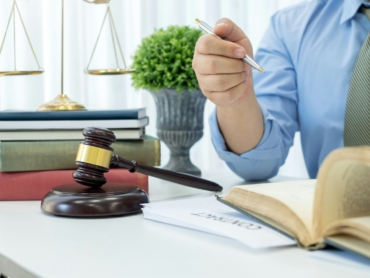 Ein Anwalt oder Richter sitzt an einem Schreibtisch mit Gesetzbüchern, einem Richterhammer und einer Waage im Hintergrund. Er hält einen Stift und liest in einem aufgeschlagenen Buch.