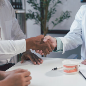 Zahnarzt schüttelt einem Patienten die Hand am Schreibtisch, daneben ein Zahnmodell – Symbol für Vertrauen und erfolgreiche Behandlung.