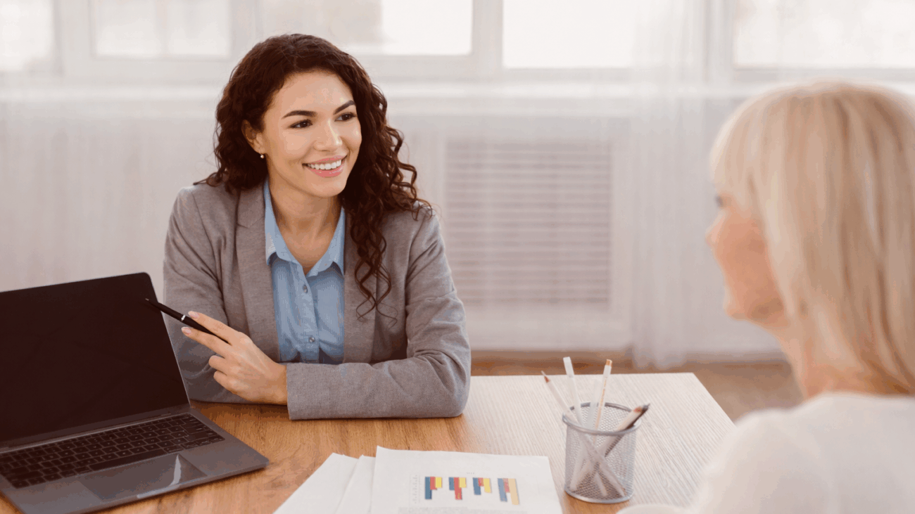 Beraterin erklärt einer Frau am Tisch ein Diagramm auf einem Dokument, Laptop daneben.