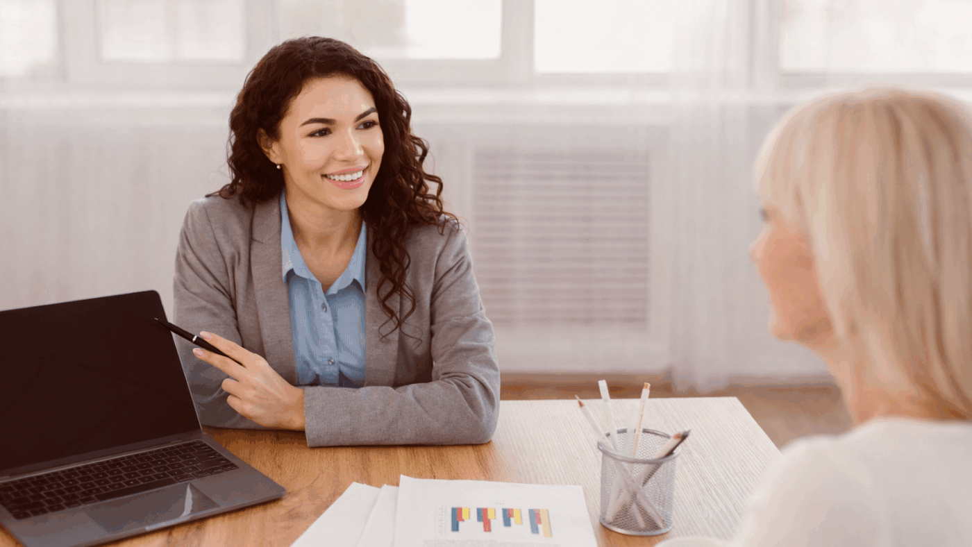 Beraterin erklärt einer Frau am Tisch ein Diagramm auf einem Dokument, Laptop daneben.