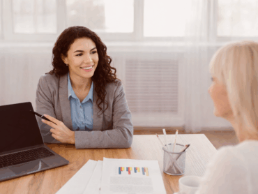 Beraterin erklärt einer Frau am Tisch ein Diagramm auf einem Dokument, Laptop daneben.