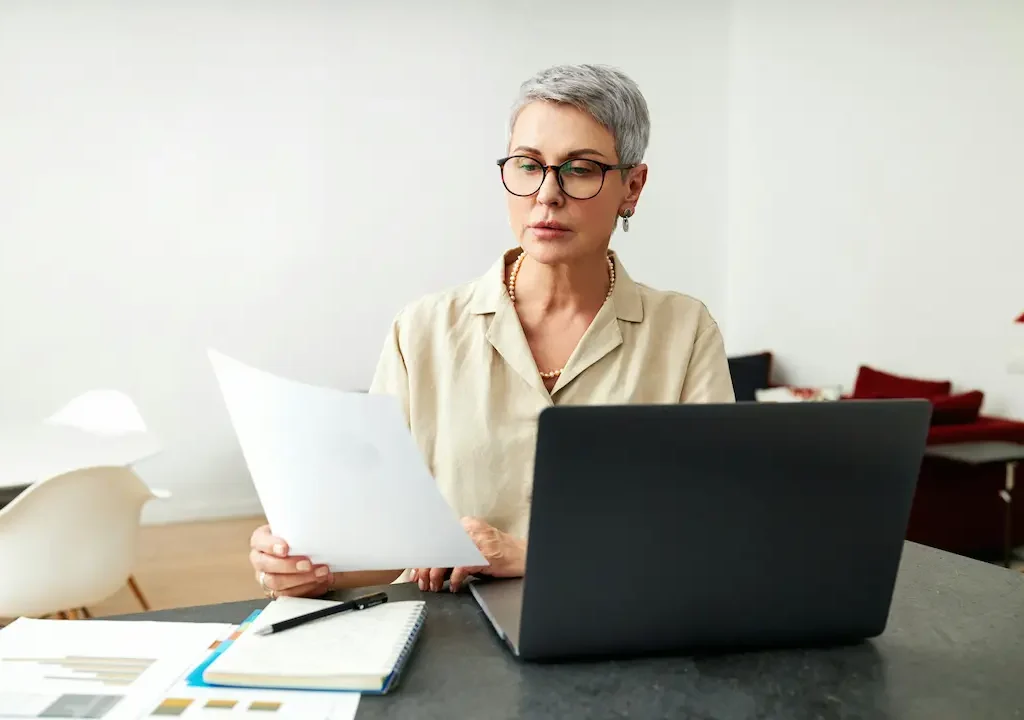 Frau mit Kurzhaarfrisur und Brille liest ein Dokument am Laptop am Schreibtisch