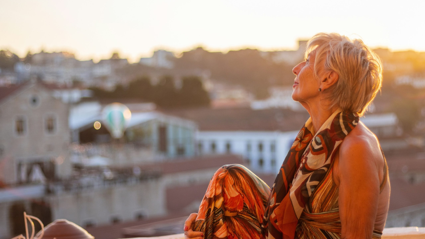 Ältere Frau sitzt entspannt auf einer Terrasse und blickt in den Sonnenuntergang über einer Stadt