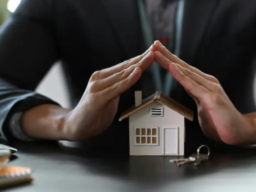 Person schützt Modellhaus mit den Händen als Symbol für Hausversicherung.
