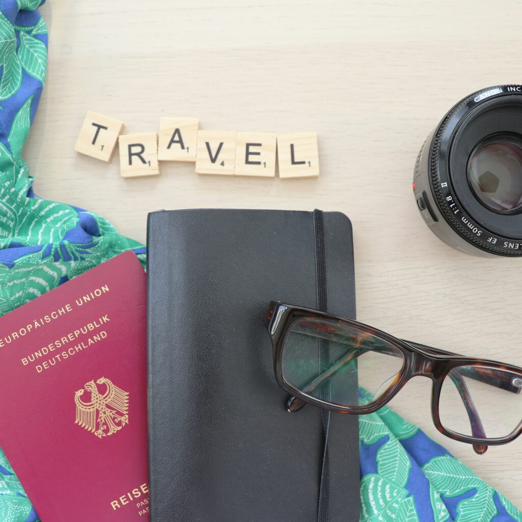 Draufsicht auf Reiseutensilien: deutscher Reisepass, schwarzes Notizbuch, Brille, Kameraobjektiv und Scrabble-Buchstaben mit dem Wort Travel
