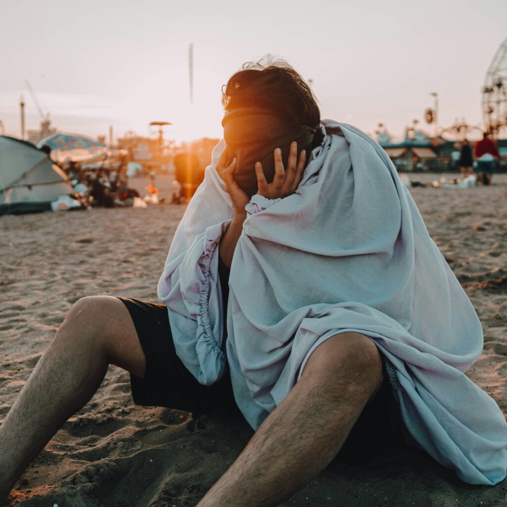 Person sitzt in eine Decke gehüllt am Sandstrand bei Sonnenuntergang, im Hintergrund ein Riesenrad und Zelte